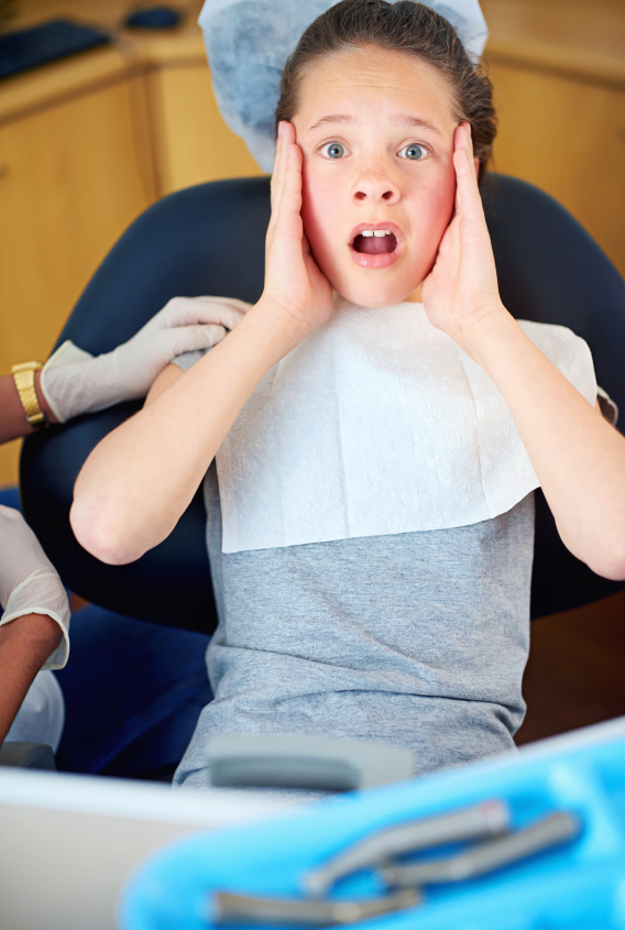 A child in a dental chair holding her head with both hands looking frightened