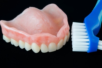 An upper acrylic denture next to a blue denture brush, sitting on a black background. An upper acrylic denture next to a blue denture brush, sitting on a black background.
