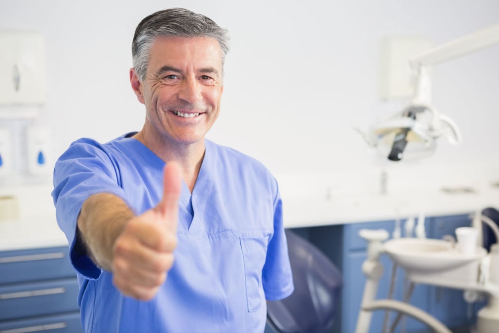 A happy dentist in a blue scrub top giving a thumbs up.