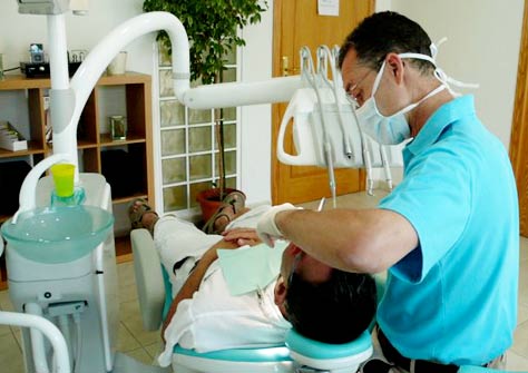 A dentist wearing a blue shirt and a mask examines a patient in a white shirt